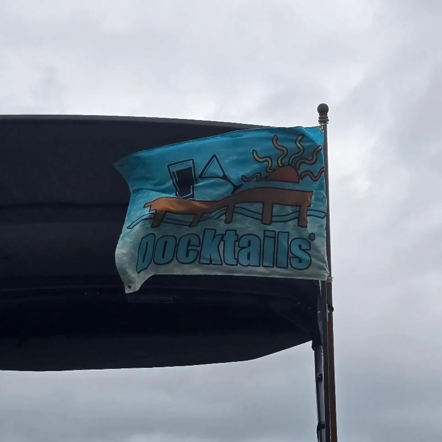 Flag flying in front of the bimini top of the boat. The flag displays the word Docktails, a sun, tilted martini glass, pint glass and a dock, set against an aqua-colored ocean backdrop.