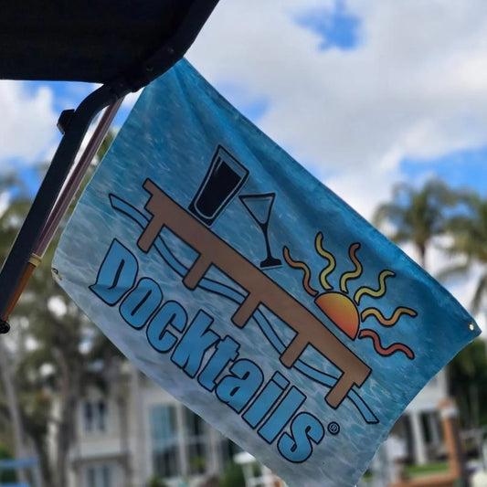 Flag hanging on an angle with an ocean background, a sun, tilted martini glass, pint glass on a dock, hanging from a boat. Background shows a blurred view of a house.