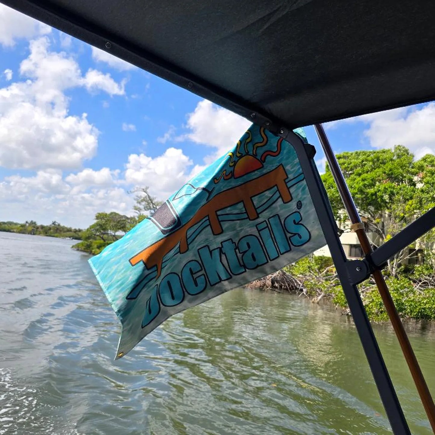 Flag flying at an angle from the bimini top of a boat, partially curled at the top, displaying the word Docktails, a sun, martini glass, pint glass and dock, set against an aqua-colored ocean backdrop. The background shows water and a building surrounded by trees.