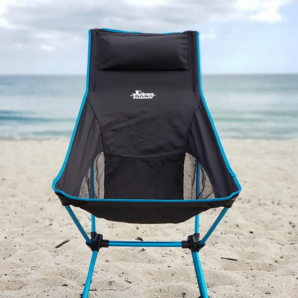 A black folding beach chair with bright blue frame and trim, featuring a built-in headrest, set on a sandy beach with the ocean and cloudy sky in the background.
