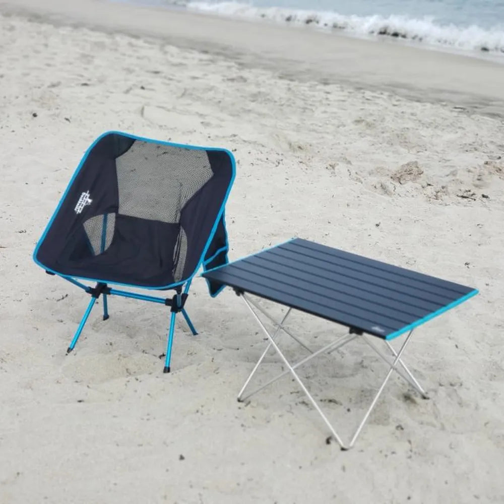 A portable camping chair and folding table set on a sandy beach with ocean waves in the background. The chair has a black mesh seat with bright blue trim and legs, and the table has a black slatted top with a blue edge trim and silver metal legs.