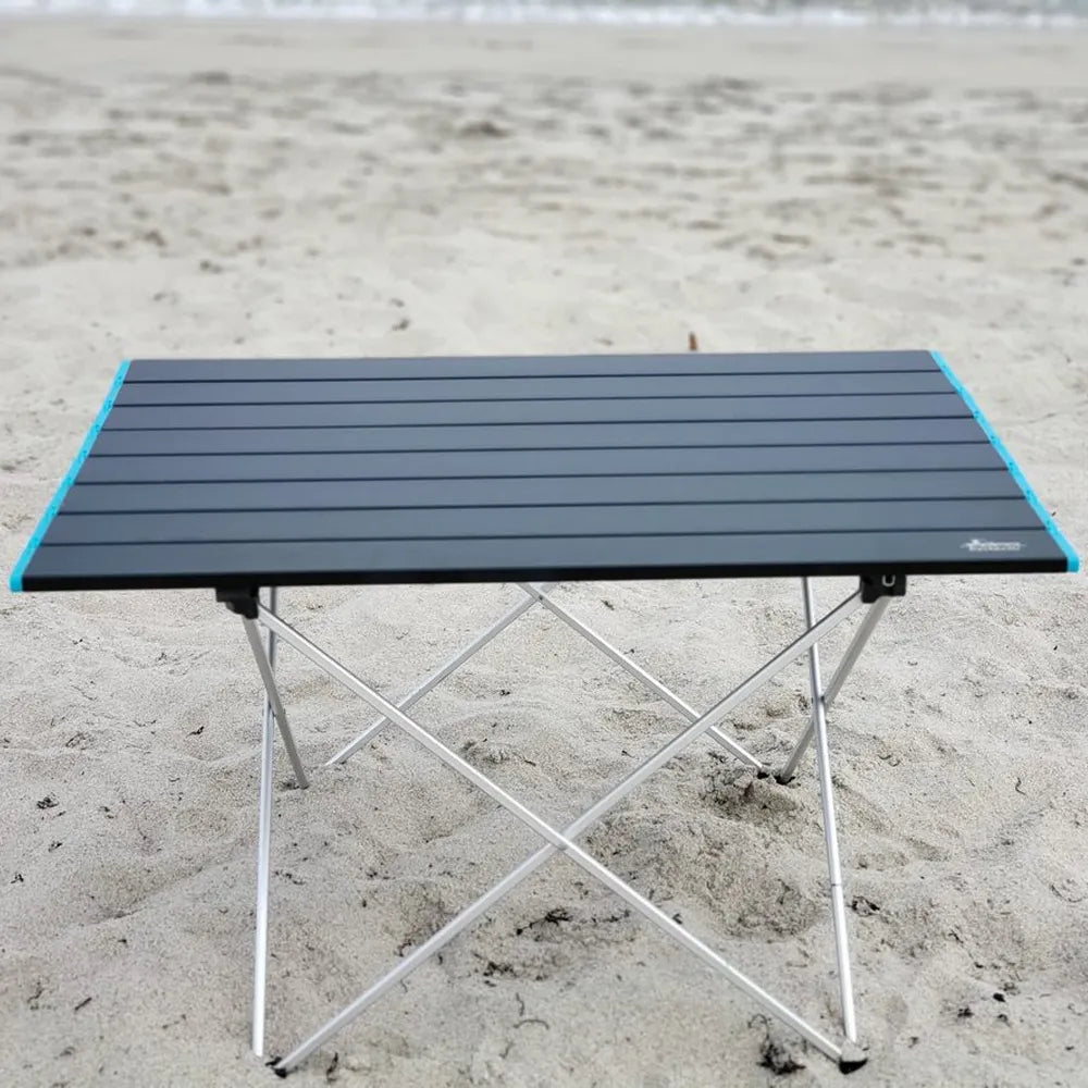 A portable folding camping table with a black slatted top and silver metal legs, set up on a sandy beach.