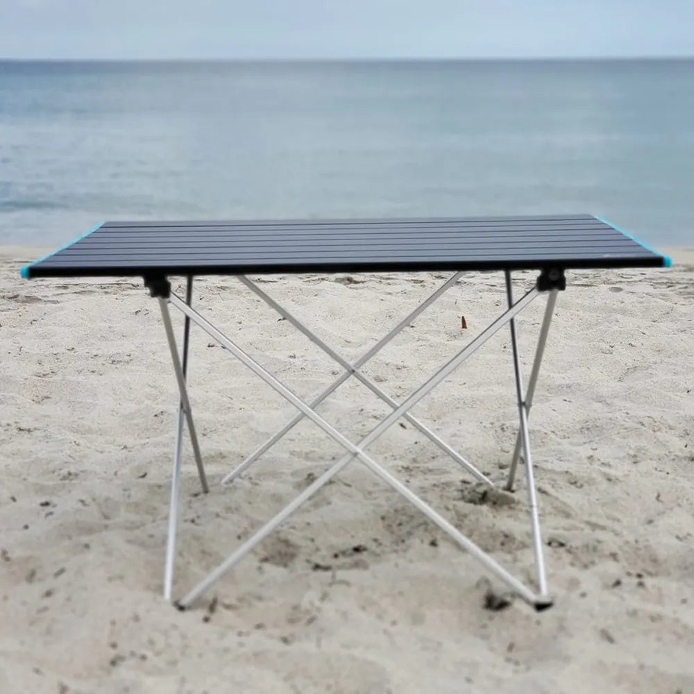 A portable folding camping table with a black slatted top and silver metal legs, set on a sandy beach with the ocean in the background.