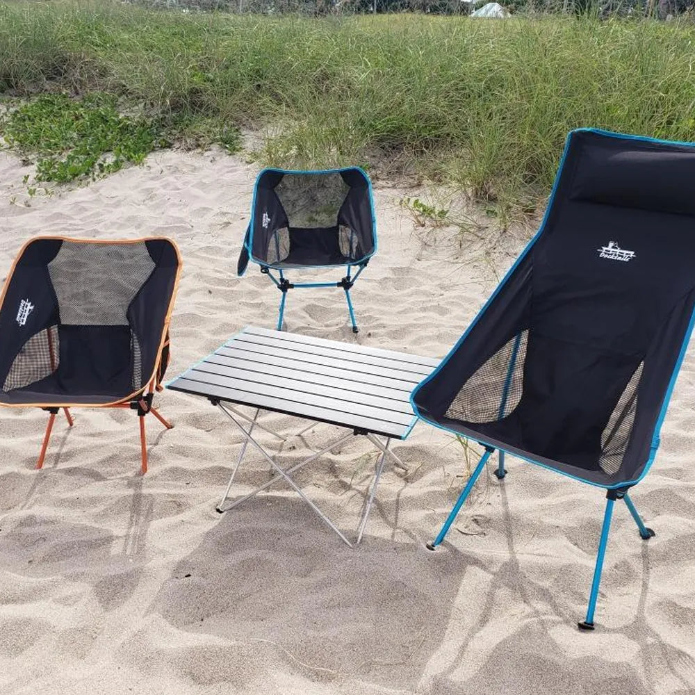 Three camping chairs and a folding table set up on sandy beach grass, with two black chairs featuring blue and orange trim and a central slatted table.