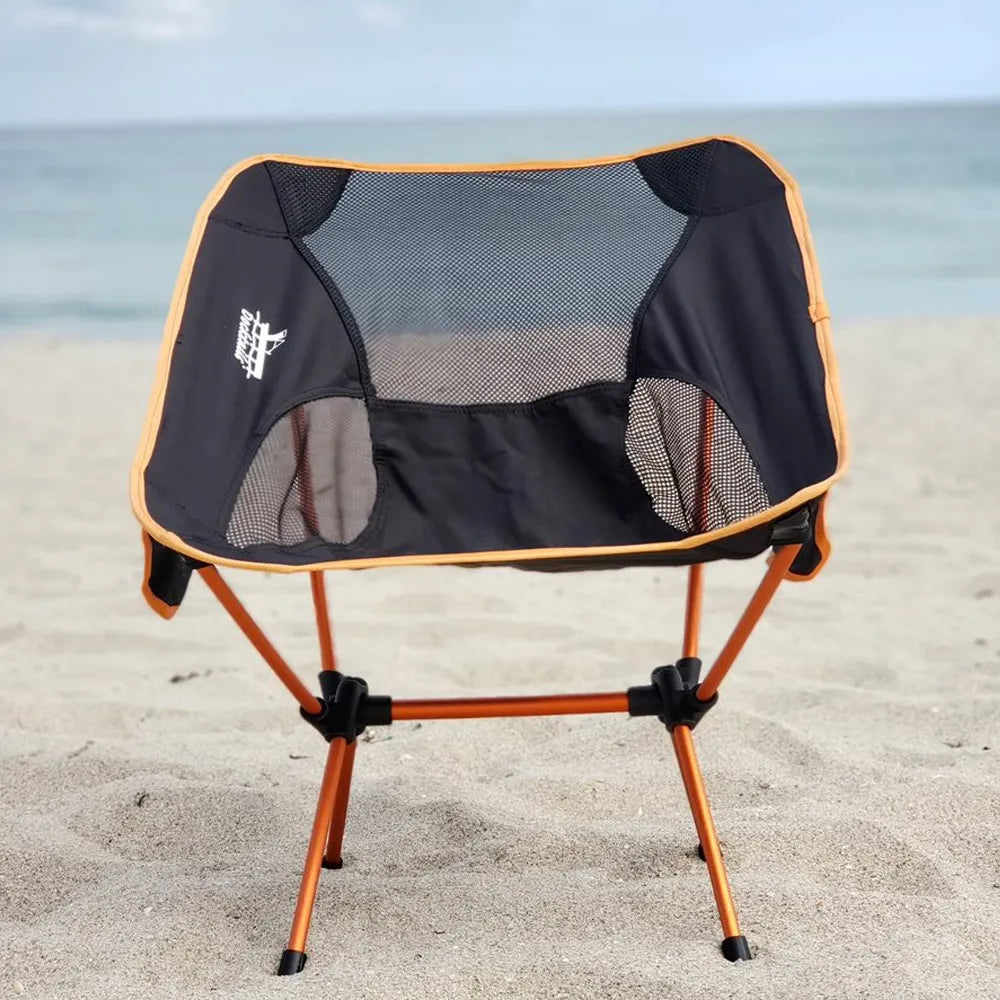 A portable folding camp chair with a black mesh seat and backrest, orange frame, and black trim, set on a sandy beach with the ocean in the background.