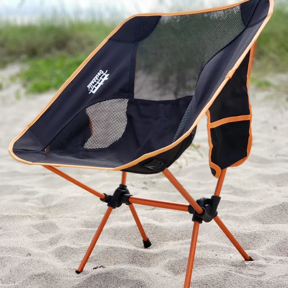A black and orange folding camp chair with mesh backrest, set on a sandy beach with blurred green grass in the background.