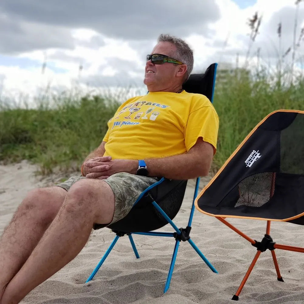 A man wearing sunglasses and a yellow t-shirt sits in a black and blue folding beach chair on a sandy beach, with another black and orange folding chair beside him and beach grass under a cloudy sky.