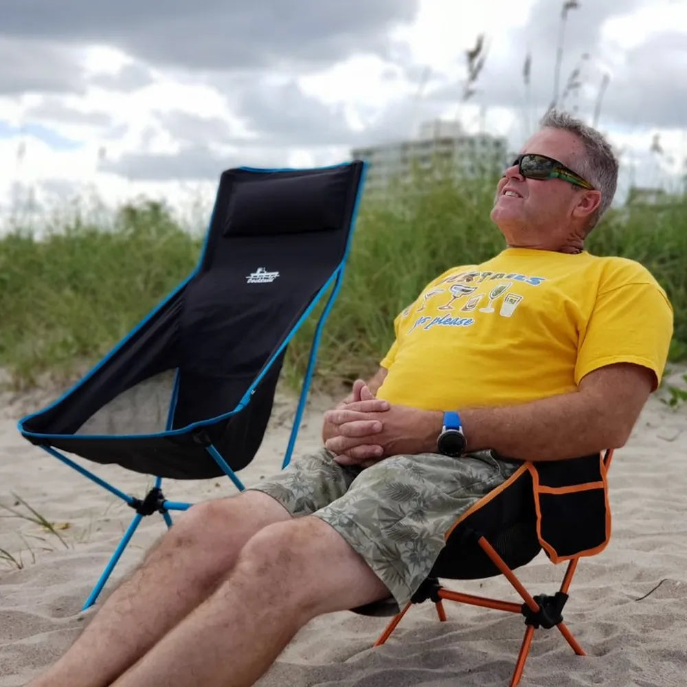 A man wearing a yellow graphic t-shirt and camouflage shorts relaxes in a black and orange folding beach chair on a sandy beach, with another black and blue folding chair beside him under a cloudy sky.