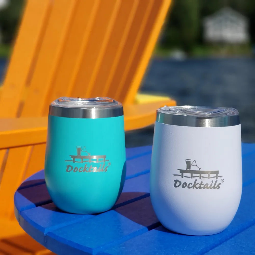 Two insulated stemless wine cups with clear lids and silver metal rims sit on a blue table outdoors. One cup is turquoise and the other is white, both engraved with a cocktail glass graphic and the word 'Docktails'.
