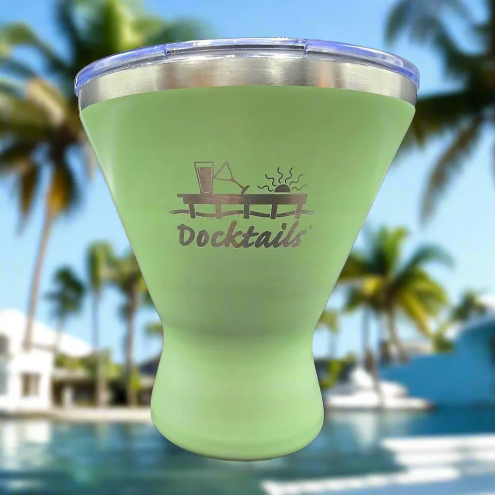 A light green tapered tumbler with a clear lid, featuring an engraved graphic of a cocktail glass and the word 'Docktails' on the front, set against a blurred tropical poolside background with palm trees.
