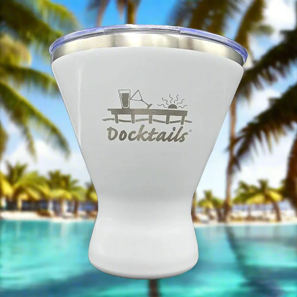 A white insulated tumbler with a clear lid and a silver metal rim, featuring an engraved 'Docktails' graphic on the front, set against a blurred tropical beach background with palm trees and a pool.