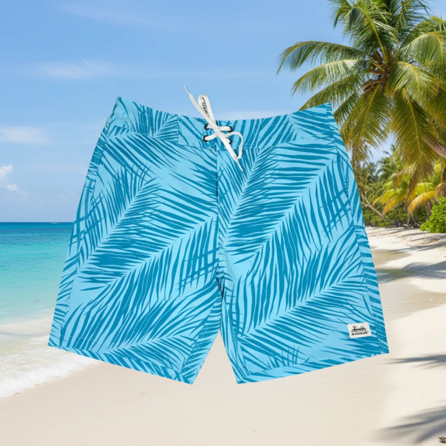 Blue tropical palm leaf print board shorts with a white drawstring, shown against a sunny beach background with palm trees and turquoise water.
