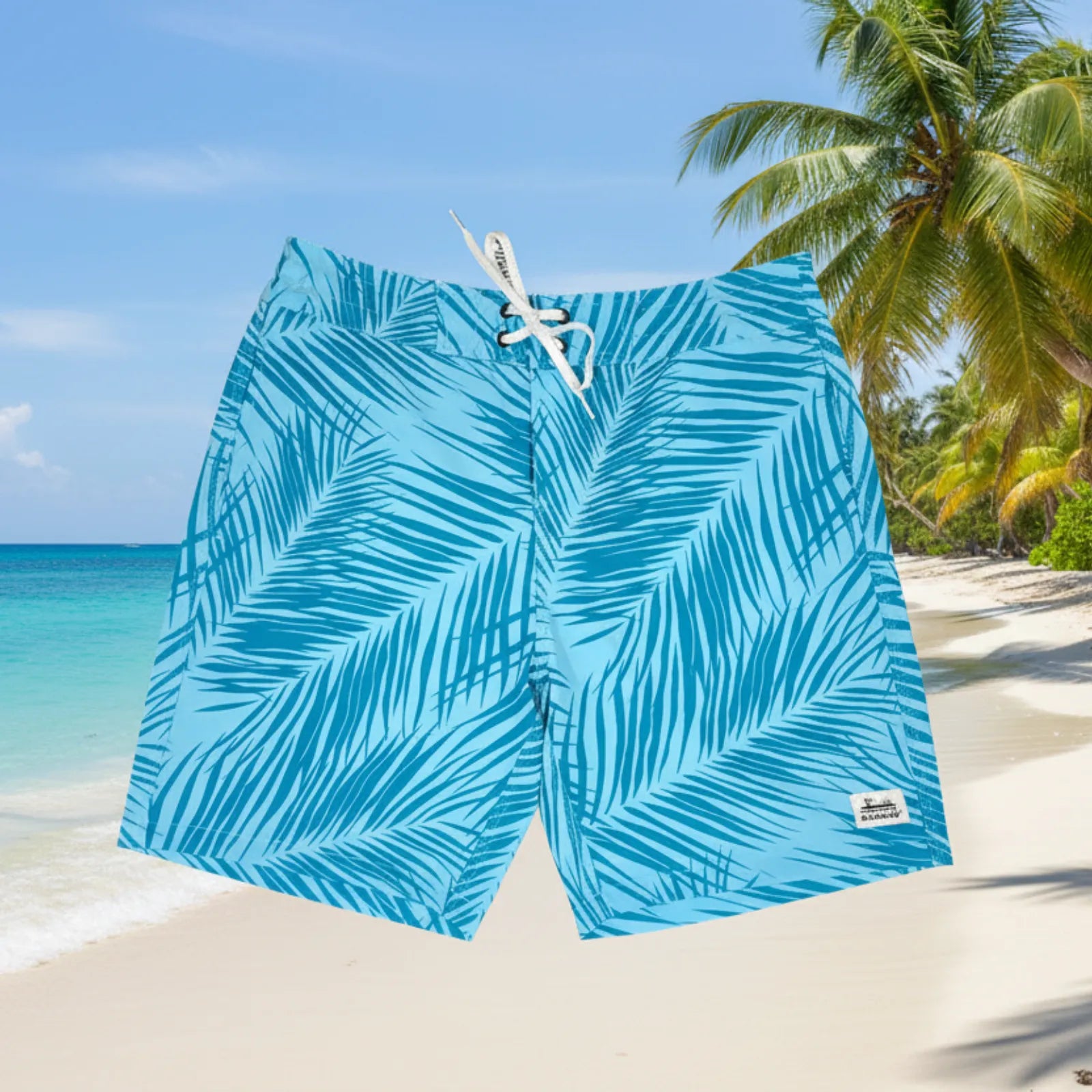 Blue tropical palm leaf print board shorts with a white drawstring, shown against a sunny beach background with palm trees and turquoise water.