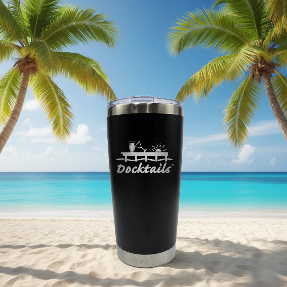 A black insulated travel tumbler with a clear lid stands on a sandy beach with turquoise ocean water and palm trees in the background. The tumbler has an engraved graphic reading 'Docktails' with a cocktail glass design.