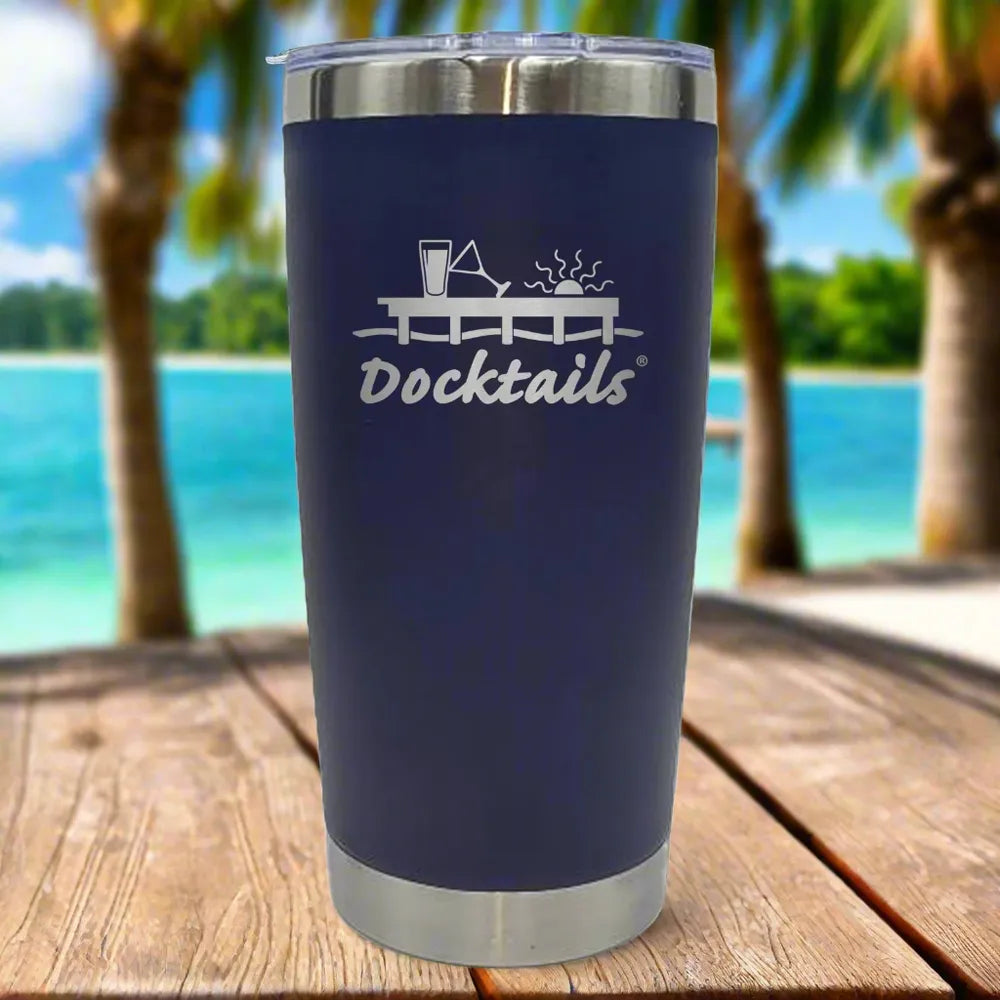 A navy blue insulated tumbler with a silver stainless steel rim and base, featuring an engraved 'Docktails' graphic on the front. The tumbler is set on a wooden table with a tropical beach background.